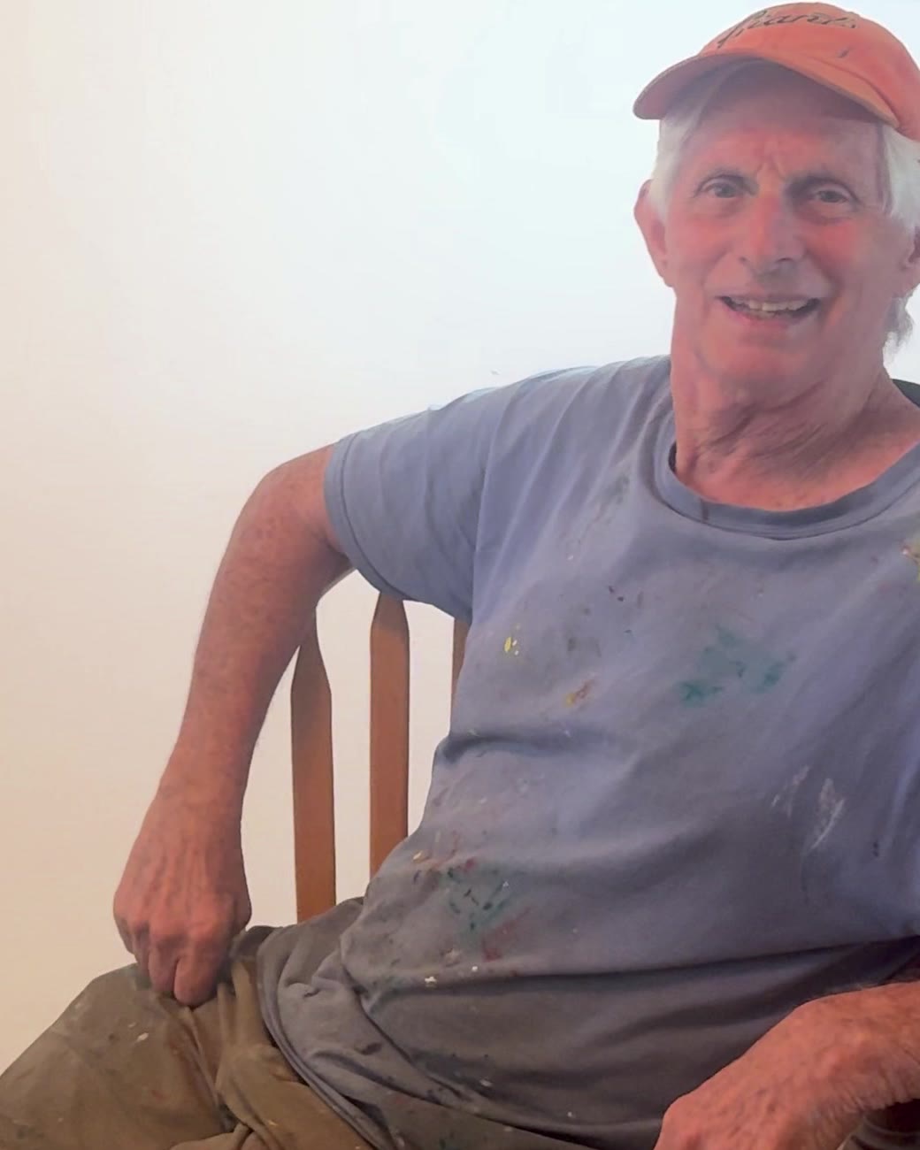 Benjamin Katz in his studio, seated in a wooden chair in a paint-flecked t-shirt and orange cap, smiling.