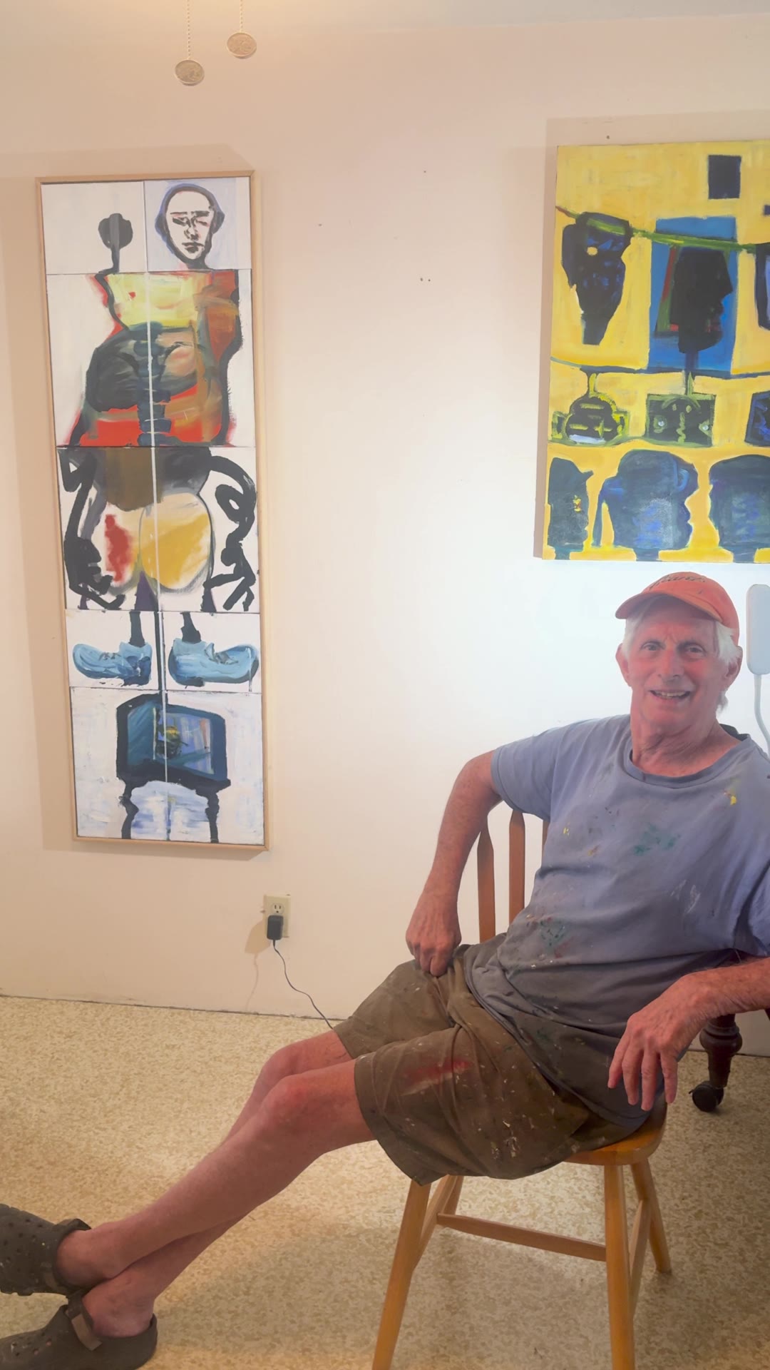 Ben Katz seated in his studio between two paintings, smiling, wearing an orange cap.